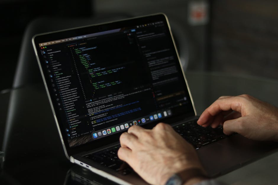 A close-up of a person's hands typing on a sleek, modern laptop with a dark theme code editor open displaying lines of programming code in green, blue, and white text. The laptop is placed on a glossy