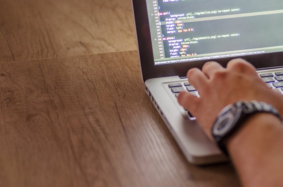 A close-up of a person's hands typing on a sleek, modern laptop with a dark theme code editor open displaying lines of programming code in green, blue, and white text. The laptop is placed on a glossy