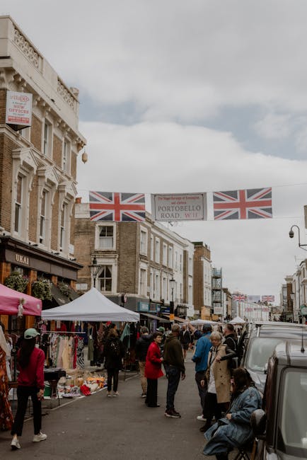 A lively street scene on Portobello Road in Notting Hill, featuring a busy outdoor market with numerous vendors and shoppers. The street is lined with historic brick buildings, some with decorative wh